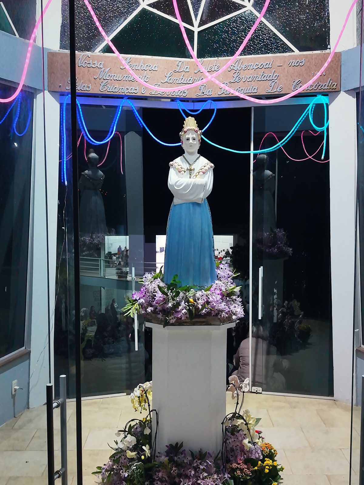 A imagem de Nossa Senhora da Salette vista de cima, dentro da capela de vidro iluminada por luzes rosa e azuis, com flores no pedestal, na noite da Romaria em Concórdia SC